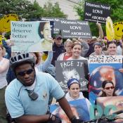 Pro-Life advocates line the march route holding signs to convey their message: WOULD YOU MARCH FOR ME [picture of fetus], WOMEN NEED LOVE NOT ABORTION, WOMEN DESERVE BETTER THAN ABORTION and FACE IT ABORTION KILLS.