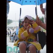 A baby sits under an umbrella in front of the Washington Monument.