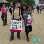 Pro-Choice activist holding demonstration sign - TERMINATE, UNWANTED, PRESIDENTS, NO TRESPASSING - The woman also has demonstration writing on her stomach - BABY FREE, [female symbol], BY CHOICE - There is also a bird drawn on her forehead.