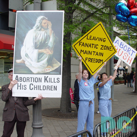 Side-By-Side: Pro-Life and Pro-Choice.  A Pro-life demonstrator holds a picture of Jesus with the message ABORTION KILLS HIS CHILDREN while Pro-Choice advocates hold signs - LEAVING FANATIC ZONE: MAINTAIN FREEDOM and ABORT BUSH.