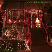 A string of bright Christmas Bells covers of porch of three Santa Claus displays. The yard is decorated with red bows, a lit Christmas tree, and seven mini wreaths.