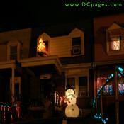 An inflatable snowman holding a candy cane waves to everyone. A reindeer and Santa can be seen on top of the porch. Lit candy canes cover the yard.