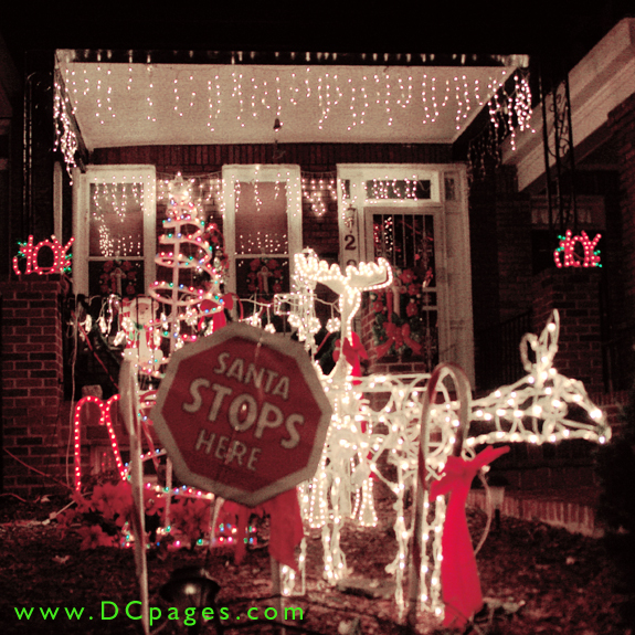 A "Santa Stops Here" sign is lit up by reindeer, candy canes, a free standing tree, Joy sign and white lights cascading from the porch. There is also man little displays on the door and windows.