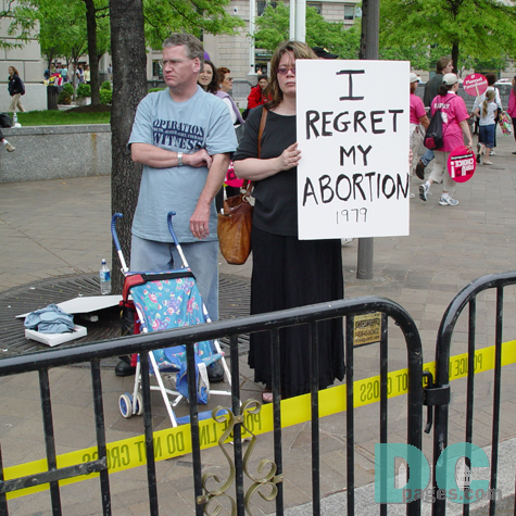 A couple with an empty baby carriage. The woman holds a sign, "I regret my Abortion."